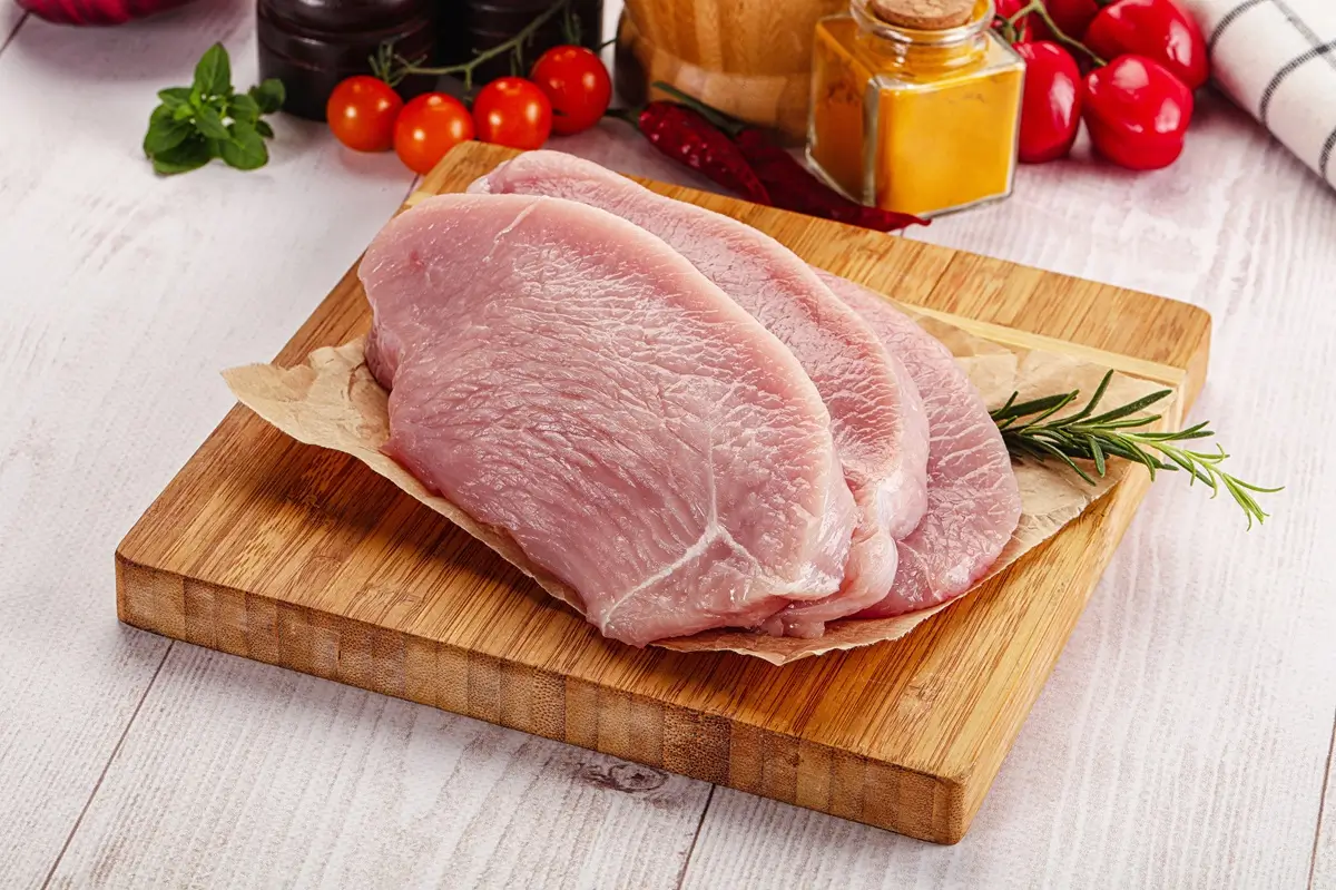 Tři plátky syrového krůtího masa na prkýnku na kuchyňském stole, v pozadí zelenina 