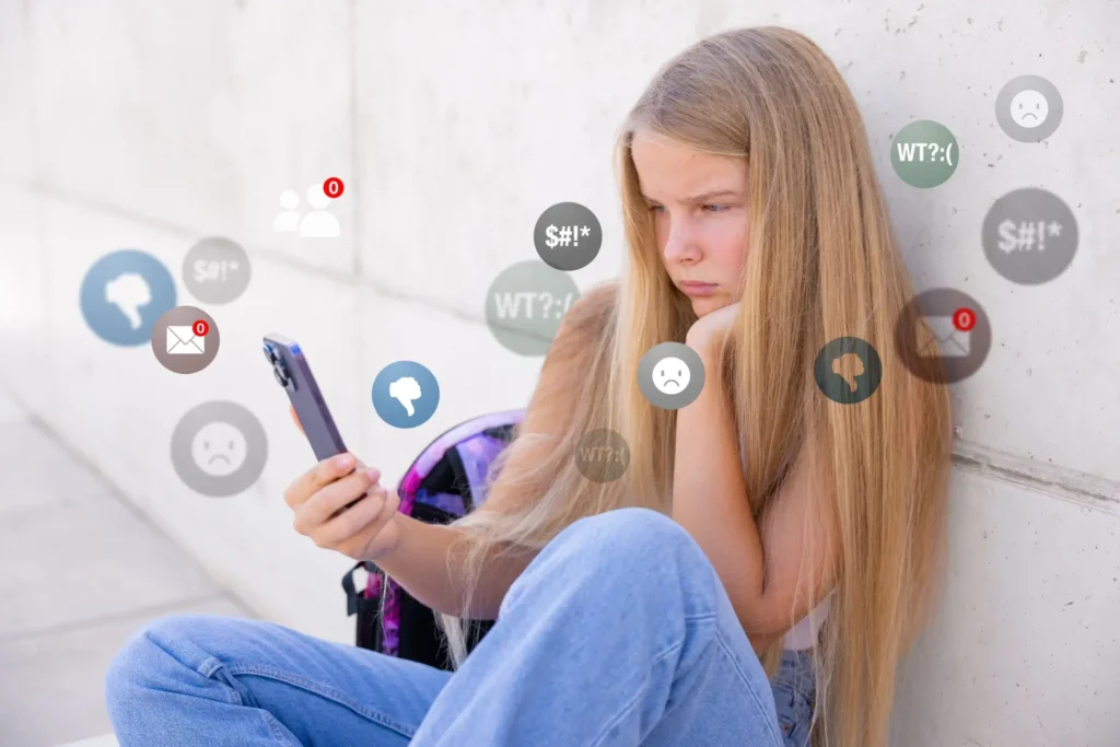Dívka sedící u zdi se zamračeným výrazem při pohledu na mobilní telefon, kolem ní ikony symbolizující online tlak a hodnocení.