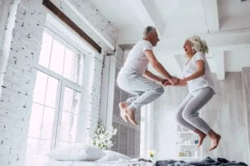Smějící se seniorský pár skáče na posteli v prosvětleném bytě