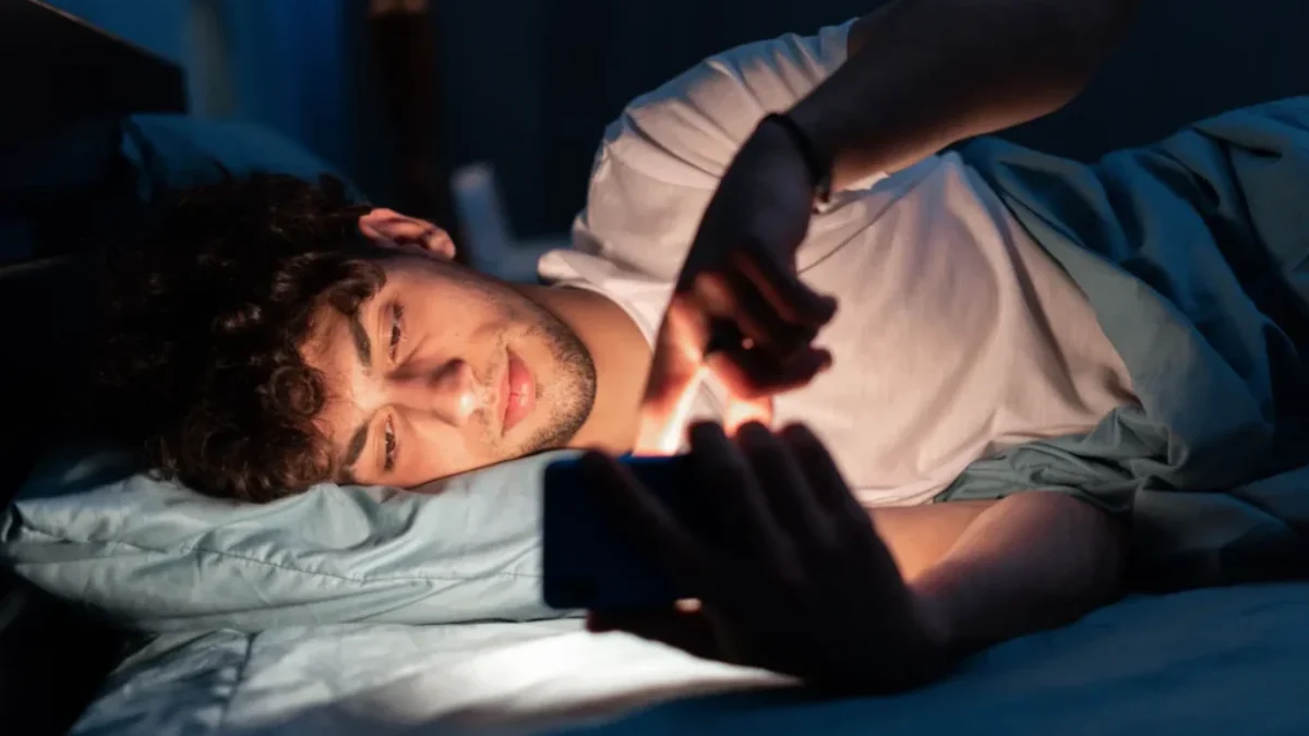 Mladý muž ležící v posteli a koukající do telefonu