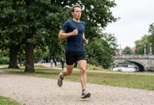 Man in a blue shirt and black shorts running along a gravel park path with trees and a river bridge in the background, mid-stride.