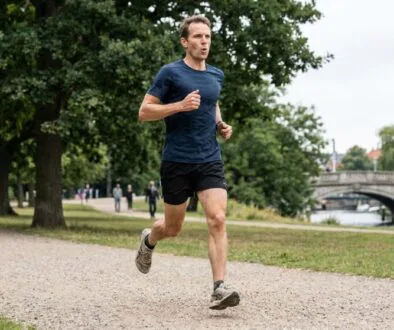 Man in a blue shirt and black shorts running along a gravel park path with trees and a river bridge in the background, mid-stride.