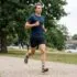 Man in a blue shirt and black shorts running along a gravel park path with trees and a river bridge in the background, mid-stride.