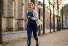 Woman jogging along a tree-lined cobblestone street with historic stone buildings in the background, smiling.