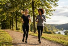 Two women in athletic wear jogging along a tree-lined trail with a river valley in the background.