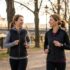 Two women jogging and smiling at each other on a sunlit park path lined with trees in autumn.